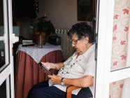 Пожилая женщина в рубашке и очках сидит на кресле и читает электронную книгу в квартире — стоковое фото