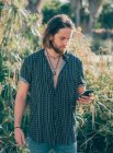 Молодой бородатый хипстер в футболке пишет смс на мобильный телефон в тропических джунглях — стоковое фото