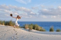 Vista lateral da fêmea em vestido branco carregando chapéu na mão correndo rápido colina arenosa na praia contra o céu azul em Tarifa, Espanha — Fotografia de Stock