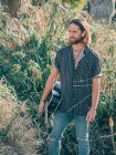 Pensive bearded hipster man standing in jungle with guitar looking away — Stock Photo