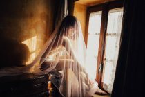 Гола чуттєва жінка вкрита прозорою білою тканиною, що сидить біля вікна на сонячному світлі — стокове фото