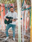 Uomo hipster barbuto suonare la chitarra acustica sotto albero decorato durante il giorno estivo — Foto stock