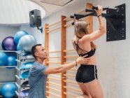 Male fitness instructor helping athletic woman to do pull ups during workout in gym — Stock Photo