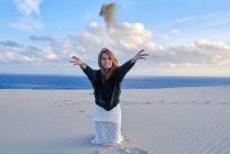Mulher alegre brincalhão jogando pilha de areia na câmera enquanto sentado na costa vazia em Tarifa, Espanha — Fotografia de Stock