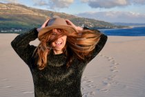 Cheerful traveling woman in hat standing in remote sandy desert on sunset looking at camera in Tarifa, Spain — Stock Photo