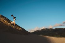 Vista laterale della femmina in abito bianco che porta il cappello in mano che corre veloce giù per la collina sabbiosa contro il cielo blu a Tarifa, Spagna — Foto stock