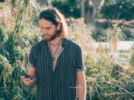 Молодой бородатый хипстер в футболке, пишет смс на мобильный телефон в тропических джунглях — стоковое фото