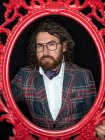 Serious impresionante hombre bien vestido con el pelo rizado y la barba posando en marco estampado rojo sobre fondo negro - foto de stock
