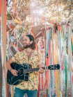 Homem hipster barbudo tocando guitarra acústica sob árvore decorada no verão — Fotografia de Stock