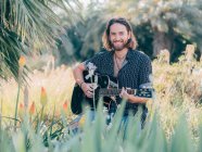 Pensive bearded hipster man sitting in meadow playing guitar looking in camera — Stock Photo