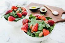 Bowls with strawberries, almonds and greenery on table with avocado and spoon of sauce on cutting board — Stock Photo
