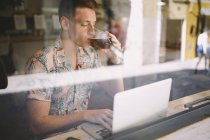 Орієнтований молодий чоловік використовує ноутбук за столом по всіх вікнах і п'є пиво в кафе — стокове фото