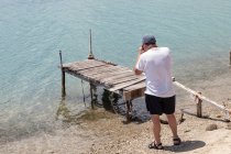 Обратный вид на неузнаваемого человека, фотографирующего серый галечный пляж с заброшенным разрушенным пирсом в кристально чистой голубой воде, Халкидики, Греция — стоковое фото