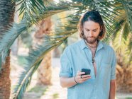 Joven barbudo hipster hombre con camisa azul mensajes de texto en el teléfono en la selva tropical - foto de stock