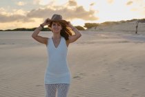 Mujer viajera alegre en sombrero de pie en el desierto de arena remota en la puesta del sol, mirando a la cámara en Tarifa, Cádiz - foto de stock