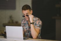 Jeune homme concentré travaillant sur ordinateur portable à la table dans un café, regardant à travers des lunettes de soleil à la caméra et souriant — Photo de stock