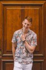 Красивый счастливый мужчина в гавайской рубашке, опирающийся на деревянную дверь, смотрящий в камеру, улыбающийся — стоковое фото