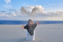 Fröhliche verspielte Frau wirft Sandhaufen in die Kamera, während sie an der leeren Küste in Tarifa, Spanien sitzt — Stockfoto