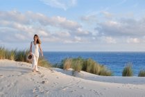 Donna allegra in abito bianco che porta il cappello in mano camminando sulla collina sabbiosa in spiaggia contro il cielo blu a Tarifa, Spagna — Foto stock