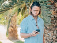 Joven barbudo hipster hombre con camisa azul mensajes de texto en el teléfono móvil en la selva tropical - foto de stock