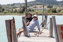Relaxing elderly man and woman in hats and sunglasses enjoying time together while sitting on pier and looking at camera in Halkidiki, Greece — Stock Photo