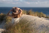 Вид сзади на женщину в шляпе, сидящую на песчаном холме и смотрящую на живописный вид на море в Тарифе, Испания — стоковое фото