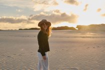 Femme voyageuse joyeuse en chapeau debout dans un désert de sable reculé au coucher du soleil, regardant la caméra à Tarifa, Espagne — Photo de stock