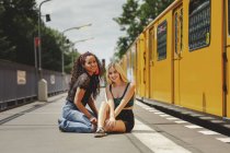 Молодые женщины сидят на железнодорожной платформе в летний день в Берлине, глядя в камеру — стоковое фото