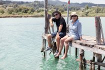 Entspannte ältere Männer und Frauen in Hüten und Sonnenbrillen genießen die Zeit zusammen, während sie auf der Seebrücke sitzen und in die Kamera schauen in Chalkidiki, Griechenland — Stockfoto