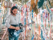 Barbuto hipster uomo suonare la chitarra acustica sotto albero decorato in estate — Foto stock
