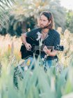 Pensive bearded hipster man sitting in meadow playing guitar looking away — Stock Photo
