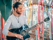 Бородатый хипстер летом играет на акустической гитаре под украшенным деревом — стоковое фото