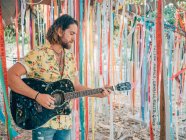 Bärtiger Hipster-Mann spielt im Sommer Akustikgitarre unter geschmücktem Baum — Stockfoto