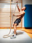 Seitenansicht eines hemdslosen muskulösen Sportlers, der beim Training in einem modernen Fitnessstudio am Seil hochklettert — Stockfoto