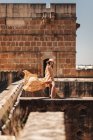 Seitenansicht einer trendigen Frau mit Hut und fliegendem Kleid, die auf einem verlassenen Gebäude an einer Ziegelmauer steht — Stockfoto
