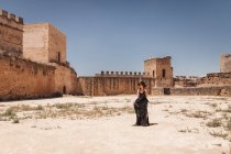 Stylish woman in black hat and long dress standing inside walls of old abandoned fortress on sunny day — Stock Photo