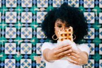 Jovem mulher em t-shirt branca em pé pela parede de azulejo colorido, comendo e mostrando waffle para câmera — Fotografia de Stock