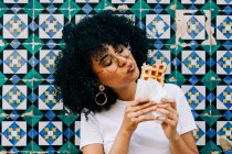 Young woman in white t-shirt standing by colorful tile wall, eating waffle — Stock Photo