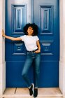 Pretty ethnic woman in white t-shirt and jeans leaning on blue door and looking at camera — Stock Photo