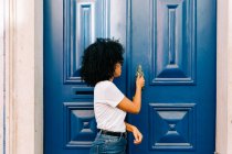 Bella donna etnica in t-shirt bianca e jeans bussare porta blu guardando altrove — Foto stock