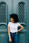 Красивая этническая женщина в белой футболке и джинсах опирается на зеленую дверь и закрытые глаза — стоковое фото