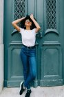 Hübsche ethnische Frau in weißem T-Shirt und Jeans an grüne Tür gelehnt — Stockfoto