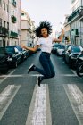 Jovem mulher afro-americana alegre em jeans e camiseta branca pulando de alegria na rua da cidade durante o dia — Fotografia de Stock