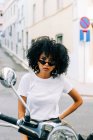 Молодая афроамериканка с черными вьющимися волосами сидит на мотоцикле и смотрит в камеру через солнцезащитные очки — стоковое фото