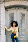 Trendige Afroamerikanerin in gelbem Crop Top und Jeans steht vor der Tür und blickt in die Kamera — Stockfoto