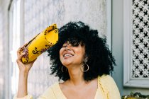 Cheerful young woman with lush curly hair and closed eyes holding fashion bag in hand and laughing — Stock Photo
