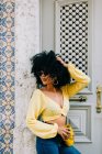 Mulher afro-americana na moda em top de cultura amarela e jeans em pé à porta e olhando para longe — Fotografia de Stock