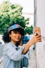 African American woman in denim jacket and colorful cap standing with smartphone and taking photo on city street — Stock Photo