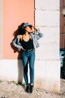Donna alla moda in jeans e canotta in piedi con gli occhi chiusi e appoggiata al muro nella giornata di sole — Foto stock