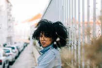 Seitenansicht einer fröhlichen Afroamerikanerin in Jeansjacke, die auf dem Bürgersteig steht und sich an Zaun lehnt — Stockfoto